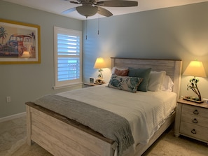 Une chambre à coucher avec un lit, deux tables de chevet avec des lampes, un ventilateur de plafond, une fenêtre avec des stores et un tableau encadré au mur.
