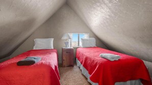 Um quarto de sótão pequeno e triangular com duas camas, uma mesa de cabeceira, um abajur e uma janela.
