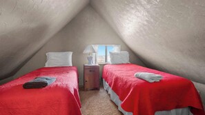 Une petite chambre mansardée de forme triangulaire, avec deux lits, une table de chevet et une fenêtre.
