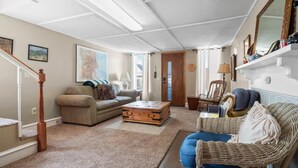 Uma sala de estar com um sofá, uma poltrona de vime, um baú de madeira e um abajur.