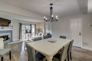 A dining area with a large table, chairs, and a chandelier. There is a fireplace with a mantel and a window with curtains.