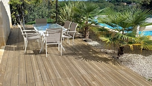 A wooden deck with a table and chairs, an umbrella, and a pool in the background. - Gîte Rêve de Caryan (Cuzance)