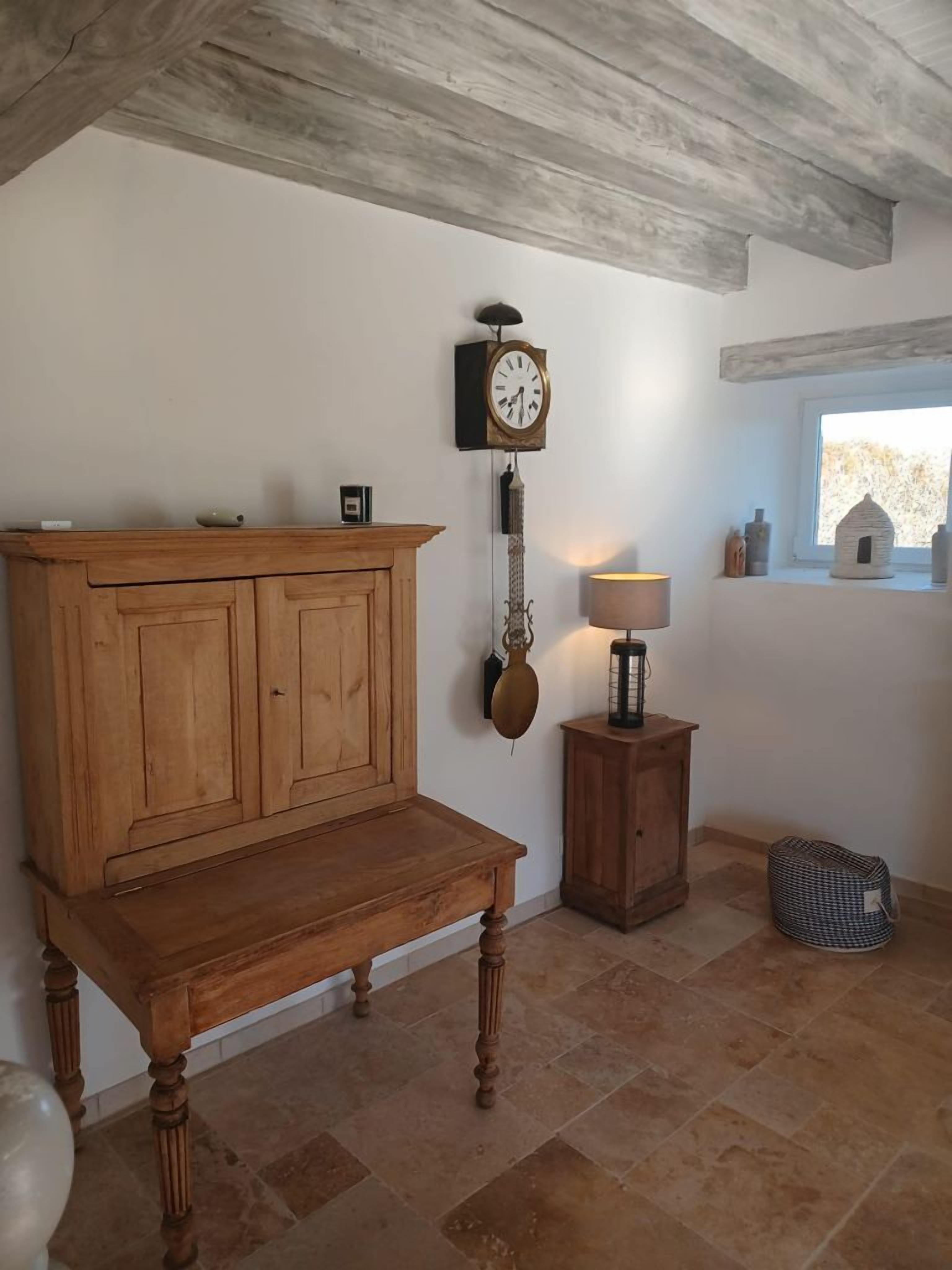 A room with a wooden cabinet, a wall-mounted clock, a small table with a lamp, and a window with a view of the outside.