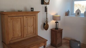 A room with a wooden cabinet, a wall-mounted clock, a small table with a lamp, and a window with a view of the outside. - Gîte Rêve de Caryan (Cuzance)