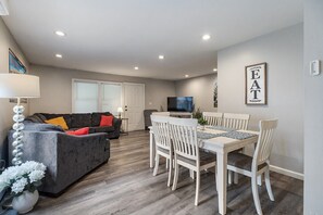 A modern living room with a grey sofa, a dining table with white chairs, a flat-screen TV, and a framed sign on the wall.