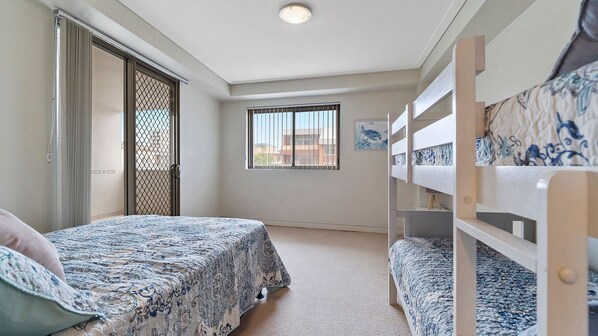 A bedroom with bunk beds, a bed with a patterned bedspread, a window with blinds, and a door with a mesh screen. - Coast on Coral Luxury Apartment - Unit 15 (The Entrance)