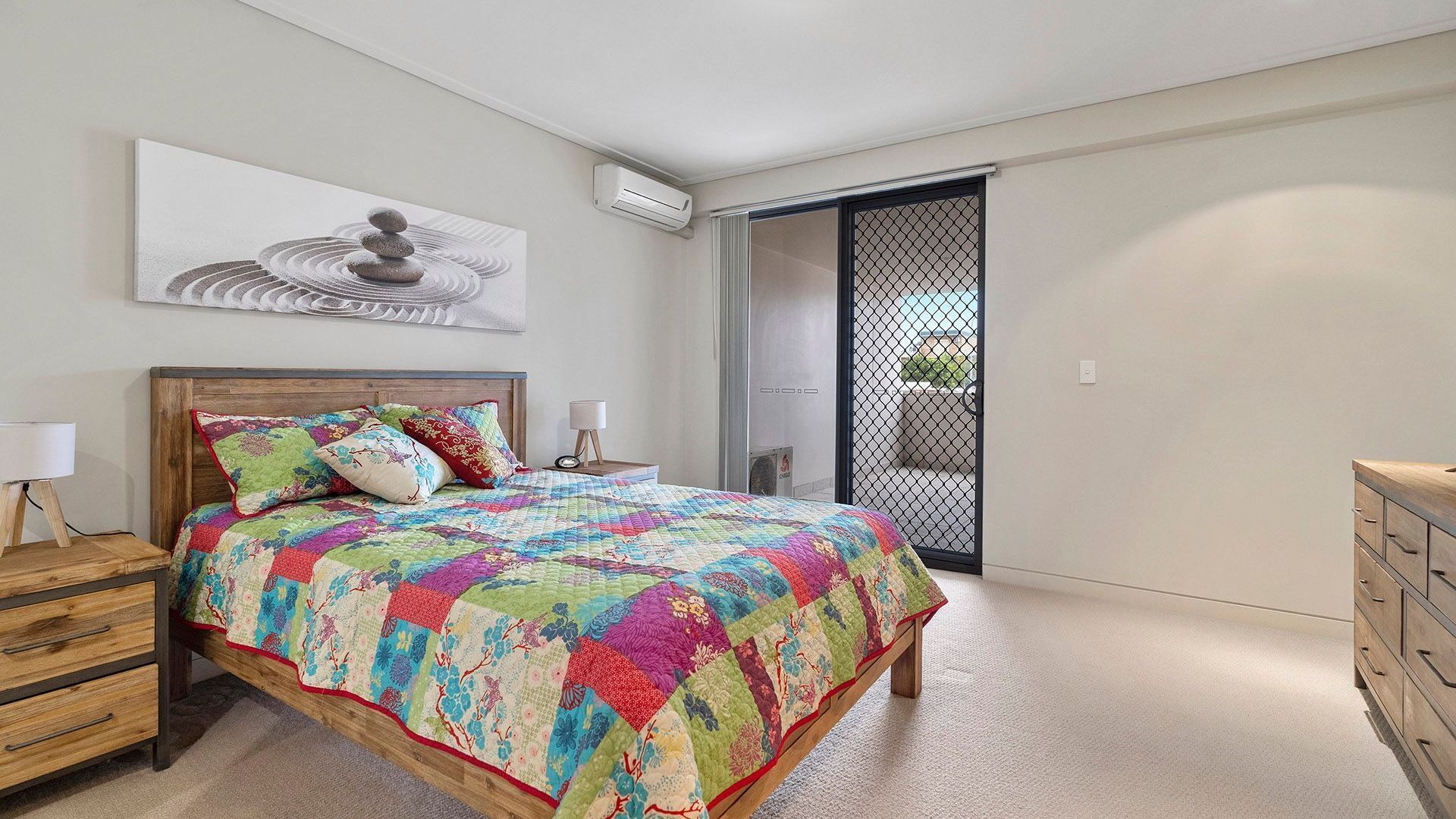 Une chambre avec un lit en bois, une courtepointe aux couleurs vives, des tables de chevet, une petite table, un climatiseur fixé au mur, une porte-fenêtre coulissante et une vue sur l’extérieur.