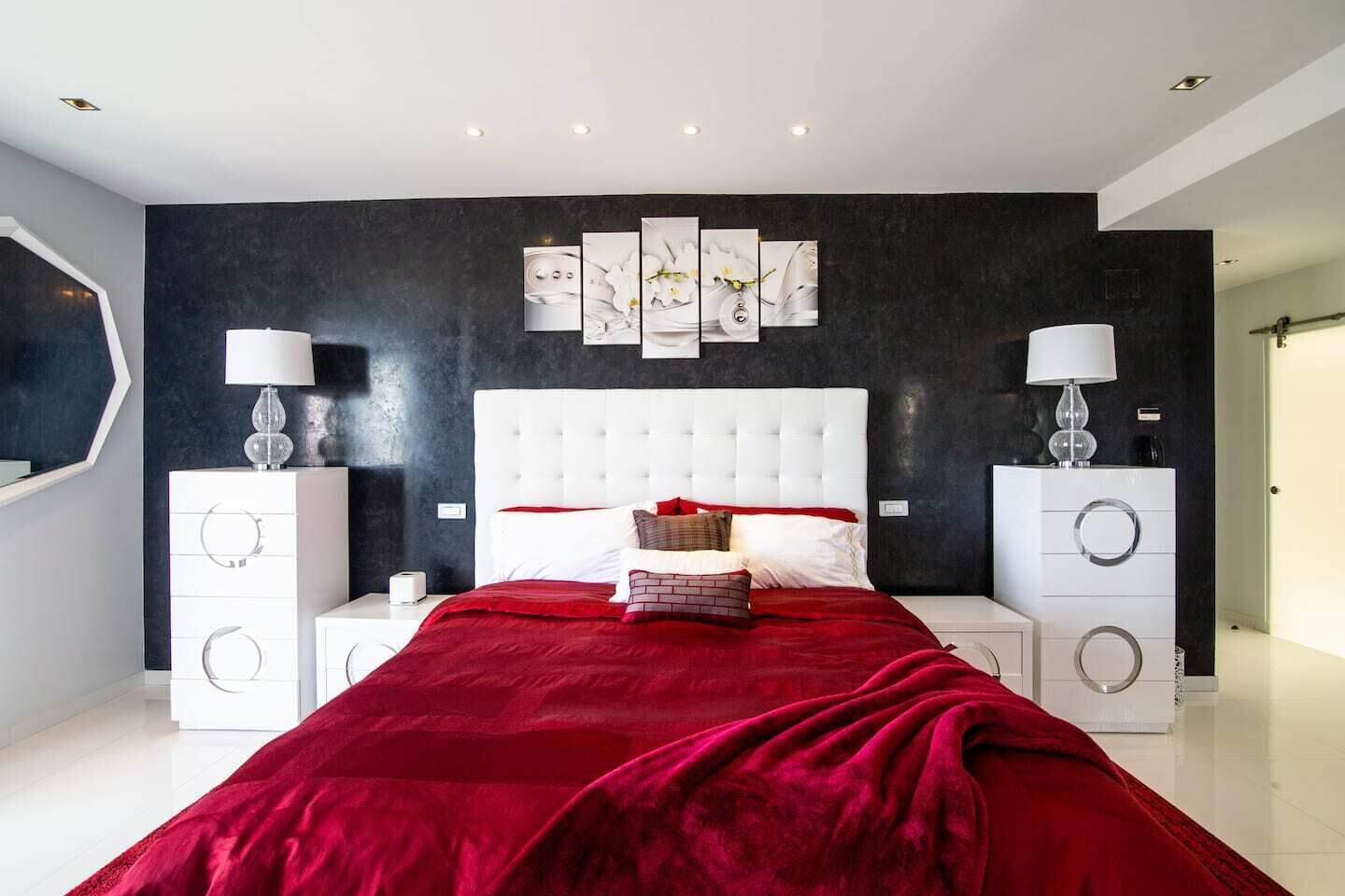 A modern bedroom with a large bed, white headboard, and red bedding. There are two bedside tables with lamps and a dark accent wall with artwork.