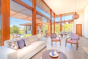 Un vaste espace de vie comprenant un grand canapé, deux fauteuils, une table basse et une table à manger avec des chaises. De grandes fenêtres offrent une vue sur l’extérieur.