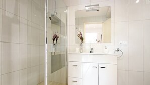 Un baño moderno con cabina de ducha de vidrio, tocador blanco con espejo y un florero con flores.