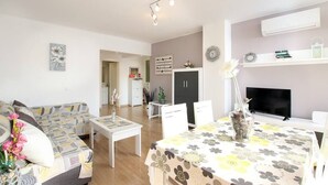 Una sala de estar moderna con sofá, mesa de centro y un comedor con una mesa dispuesta para dos personas. Hay un televisor sobre un mueble bajo, una chimenea y una estantería con objetos decorativos.