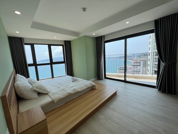 A modern bedroom with a large bed, a wooden bedside table, and a view of the sea through large windows.