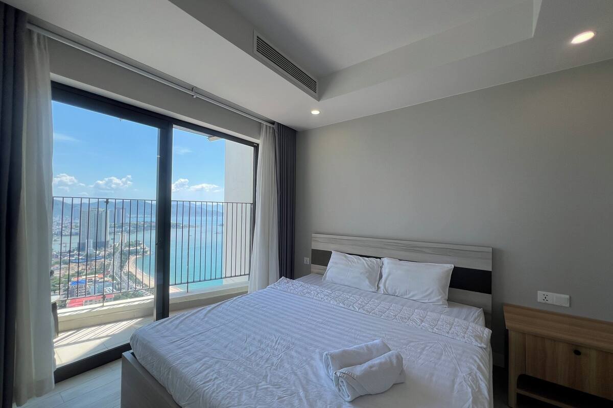Een hotelkamer met een groot bed, een houten nachtkastje en een uitzicht op een stadsgezicht vanaf een balkon.
