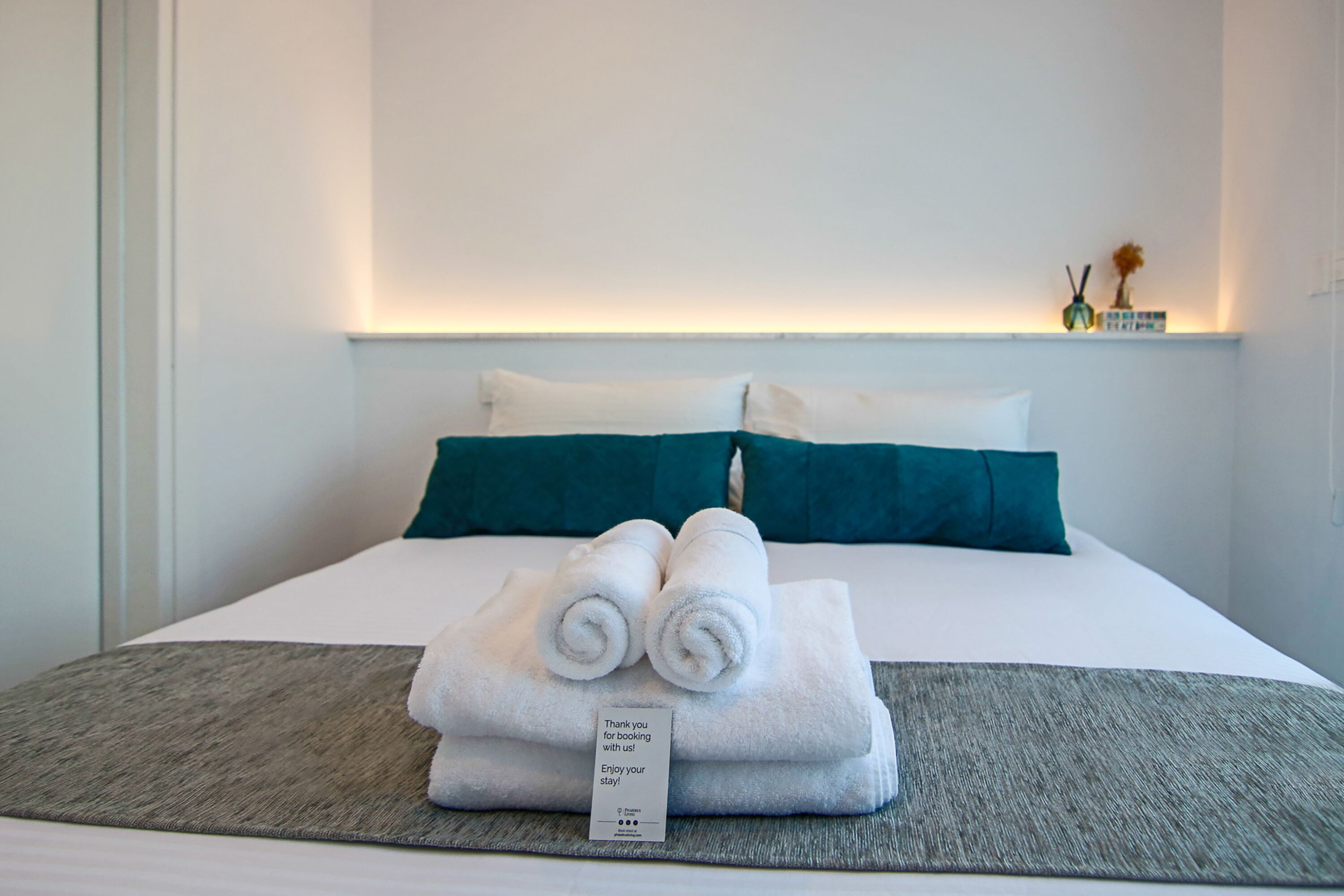 Ein ordentlich bezogenes Bett mit weißen Leinen und Türkis-Kissen, ein gefaltetes Handtuch auf dem Bett und ein kleines Schild mit einer Botschaft.