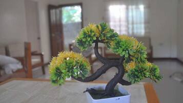 Een pot met een bonsai-boom, herkenbaar aan de dikke stam en vele takken, staat op een tafel in een kamer met een bed en een raam op de achtergrond.