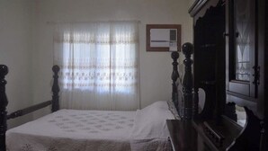 Un dormitorio con cama, un armario de madera, una ventana con cortinas translúcidas y un equipo de aire acondicionado instalado en la pared.