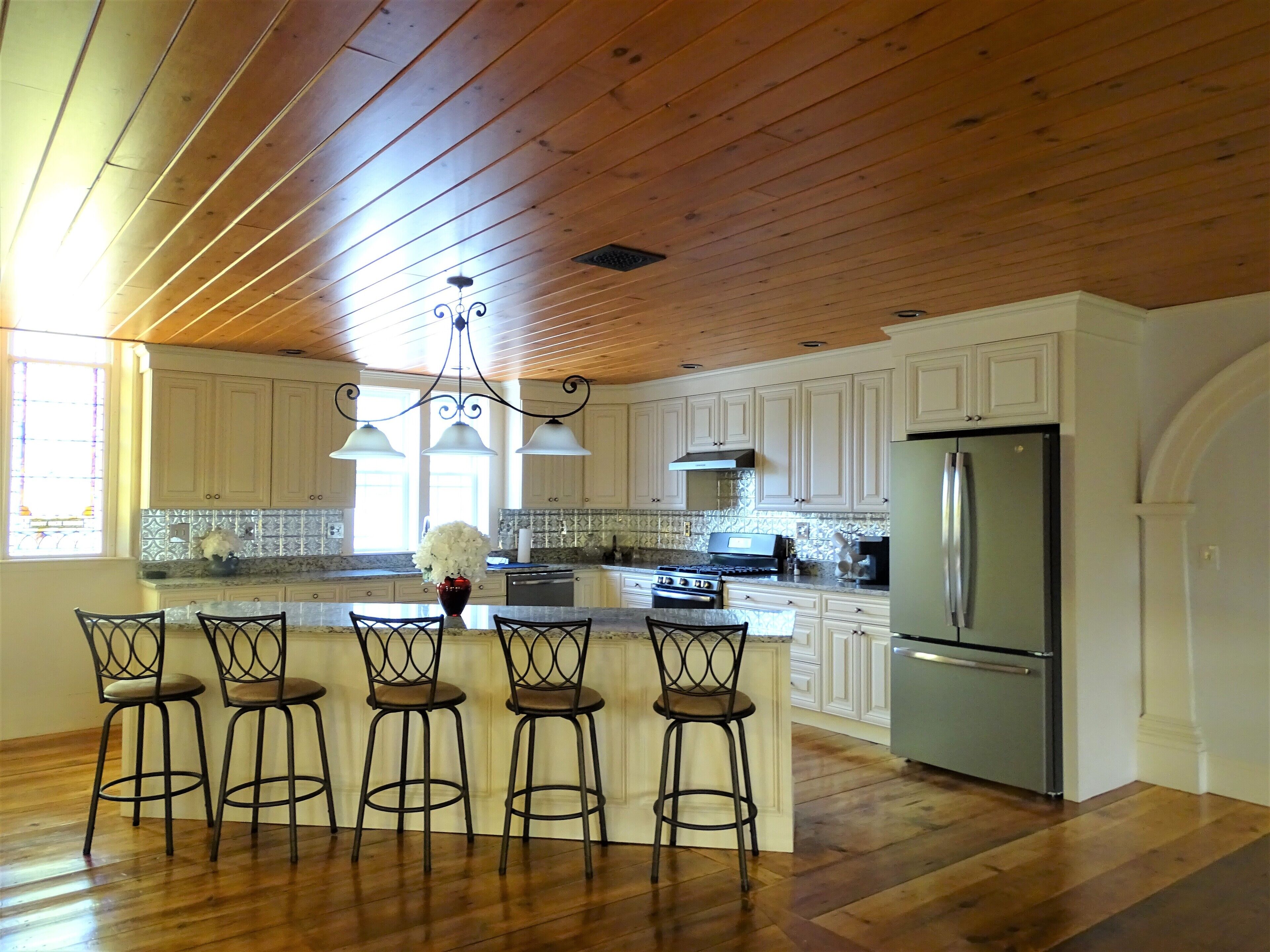 A kitchen with wooden ceiling, white cabinets, a stainless steel refrigerator, and a bar area with stools.