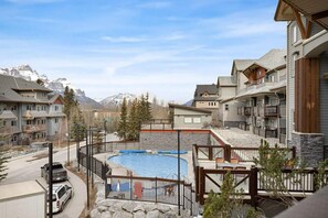A residential area with a swimming pool surrounded by houses, a road with parked cars, and mountains in the background.