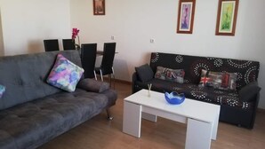 Uma sala de estar com um sofá cinza, uma mesa de centro branca e um sofá preto com almofadas estampadas. Há duas imagens emolduradas na parede.