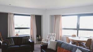 En moderne stue med en sort lædersofa, en polstret lænestol og et spisebord med stole. Store vinduer med gardiner giver naturligt lys og udsigt.
