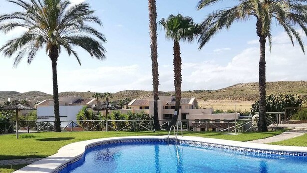 A swimming pool with clear blue water, surrounded by palm trees and a building in the background.