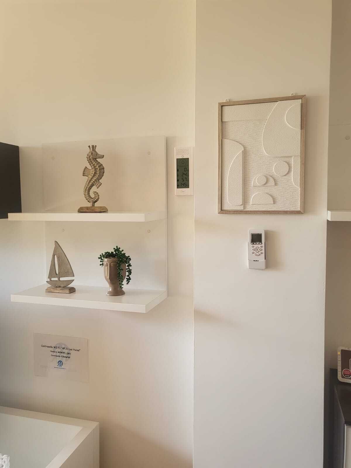 A minimalist room with white walls, a framed abstract artwork, a digital thermostat, and a small potted plant on a shelf.