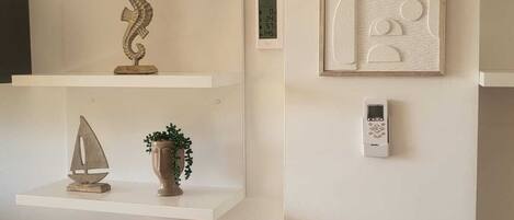 A minimalist room with white walls, a framed abstract artwork, a digital thermostat, and a small potted plant on a shelf.