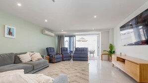 Un salon moderne avec un canapé gris, deux fauteuils bleus, un meuble TV en bois et une grande fenêtre donnant sur des maisons.