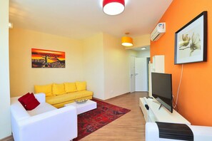 A modern living room with a yellow sofa, white armchair, and a red pillow. A flat-screen TV is on a white stand, and a painting is on the wall. - Rio Spot Homes T037 Ocean View (Rio de Janeiro)