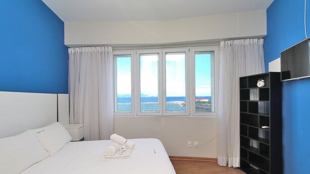 A hotel room with a bed, a nightstand, a TV, and a view of the sea through large windows. - Rio Spot Homes T037 Ocean View (Rio de Janeiro)
