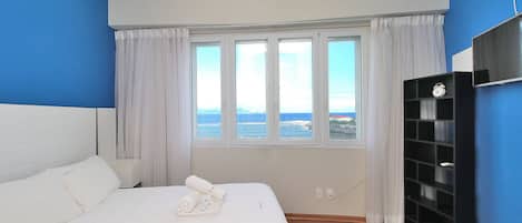 Habitación de hotel con cama, mesita de noche, televisor y vista al mar a través de amplias ventanas.