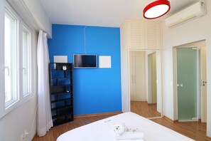A bedroom with a blue wall, a bed with a white comforter, a TV, a shelf with a clock, and a door with a mirror. - Rio Spot Homes T037 Ocean View (Rio de Janeiro)