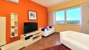 A living room with a flat-screen TV, white sofa, orange walls, and a large window with a view. - Rio Spot Homes T037 Ocean View (Rio de Janeiro)