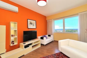 A living room with a flat-screen TV, white sofa, orange walls, and a large window with a view. - Rio Spot Homes T037 Ocean View (Rio de Janeiro)
