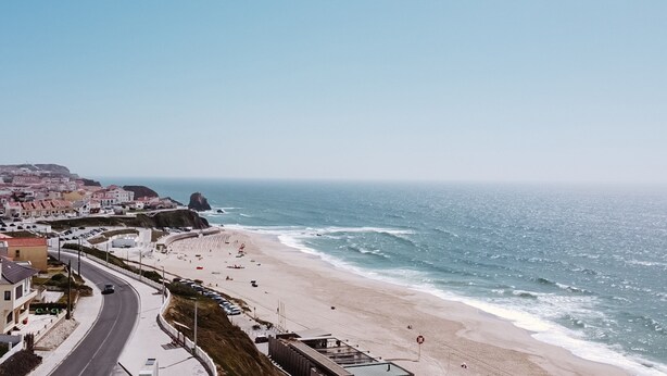 Una strada costiera che conduce a una spiaggia, con edifici e un promontorio roccioso all'orizzonte.