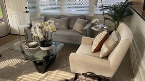 Ein Wohnzimmer mit einem Glastisch, einem beigefarbenen Sessel und einem grauen Sofa. Eine weiße Tür und ein Fenster mit Jalousien sind vorhanden.