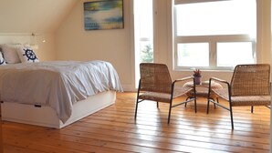 A bedroom with a bed, two wicker chairs, a painting, and a large window.