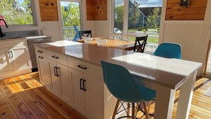 A modern kitchen with wooden flooring, white cabinetry, a central island with bar stools, pendant lighting, and large windows offering views of the outdoors.