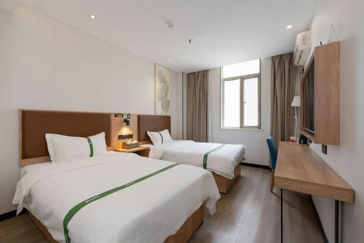 A hotel room with two beds, a desk, a lamp, and a window.