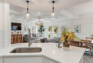 A spacious kitchen with a central island, a dining area with wooden chairs, and a living room with a gray sofa and decorative pillows.