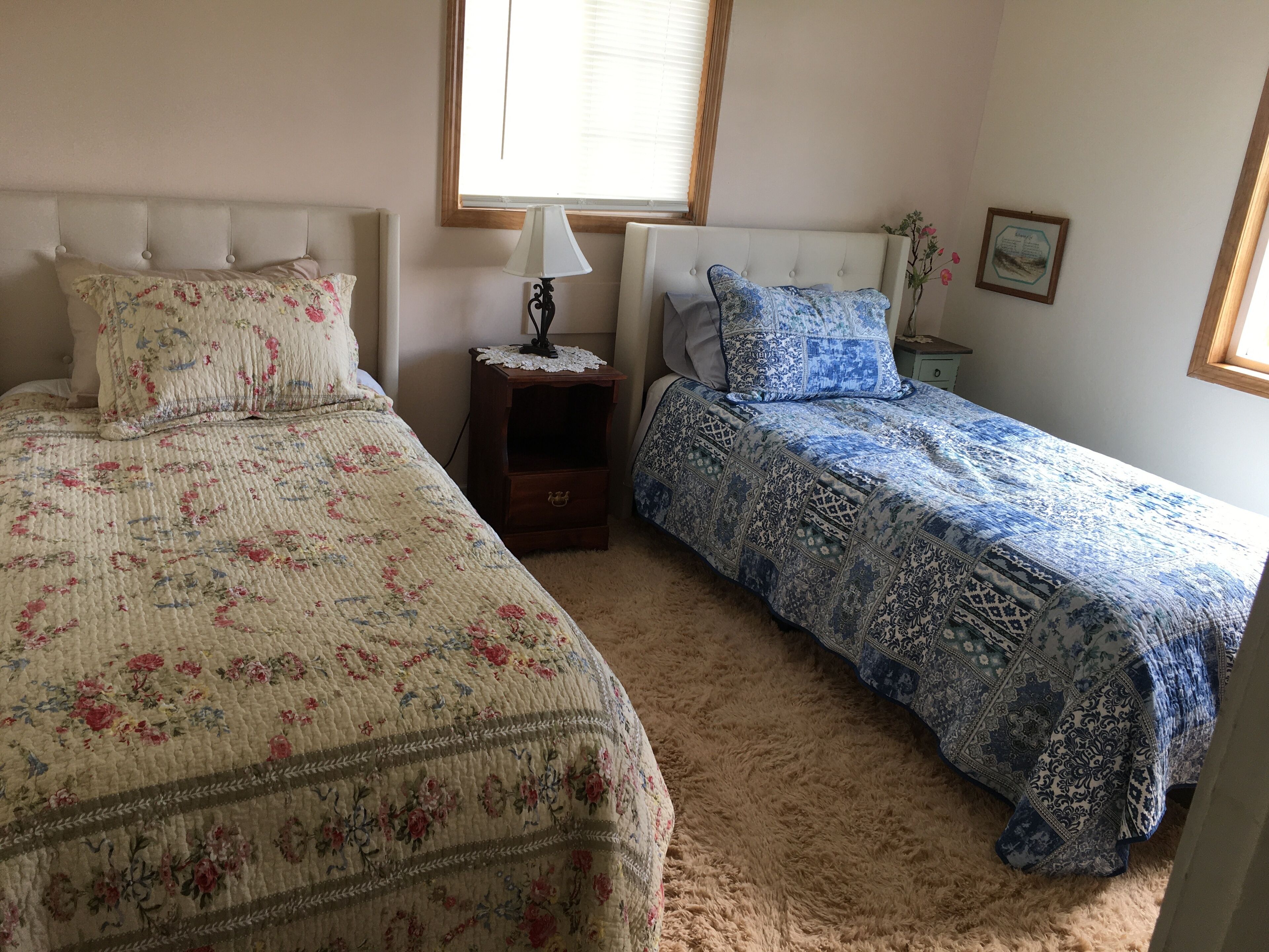A bedroom with two beds, each with a distinct floral and blue patterned bedspread. There is a nightstand with a lamp between the beds, and a framed picture on the wall.