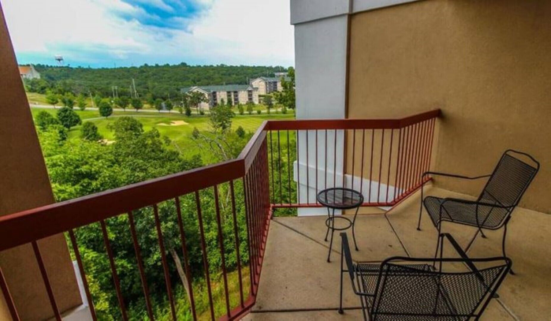 Ein Balkon mit roter Brüstung, zwei Metallstühlen und einem kleinen Tisch. Im Hintergrund ist ein Blick auf einen Golfplatz und Gebäude zu sehen.