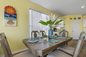 Un coin repas avec une table en bois dressée pour quatre personnes, entourée de chaises. Une grande plante en pot est posée sur la table, et un élément décoratif orne le mur au-dessus.