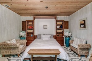 Un dormitorio con una cama grande, dos sillas de mimbre, un banco de madera y estanterías empotradas con cestas y objetos decorativos.