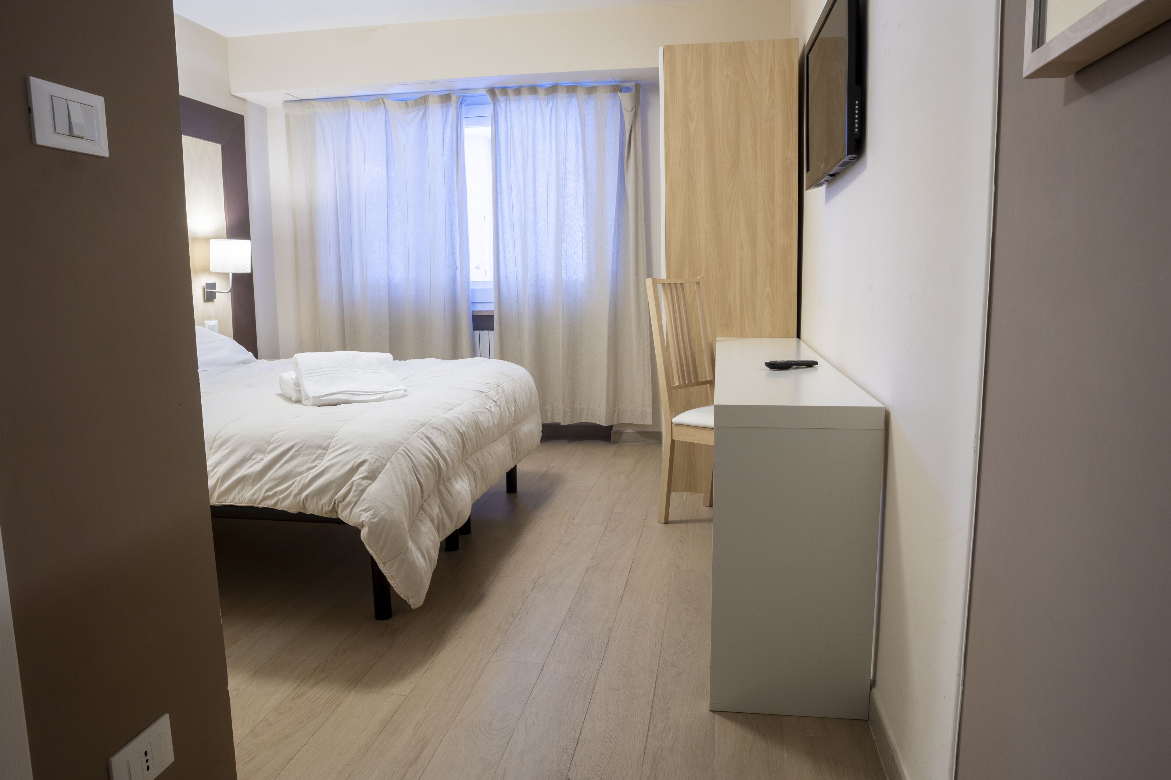 Ein Hotelzimmer mit Bett, Schreibtisch mit Stuhl, Fernseher und einem Fenster mit Vorhängen.