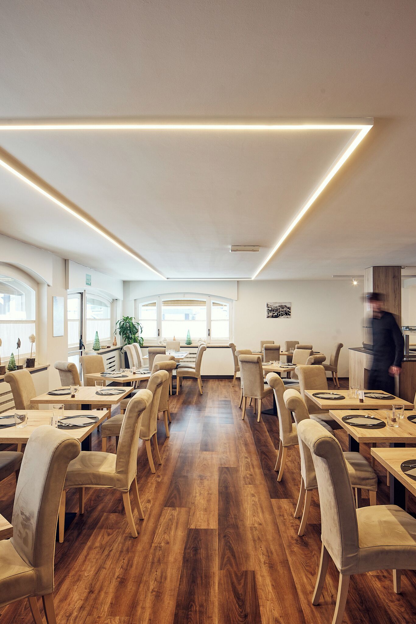 Un ristorante moderno con pavimento in legno, sedie imbottite color beige e un lungo tavolo con un cameriere che si muove.