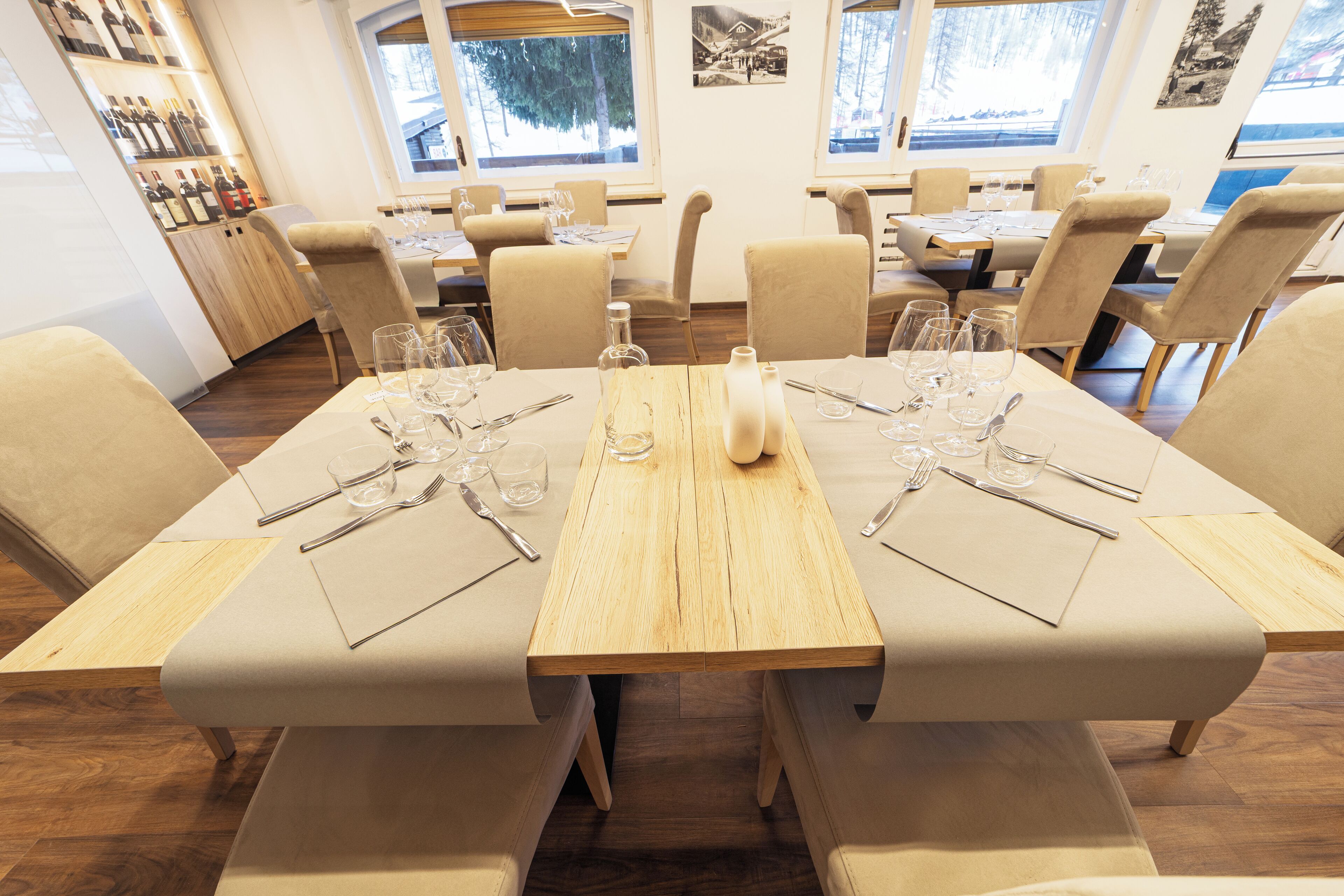Un restaurante con mesas de madera y sillas tapizadas en color beige, listo para el servicio con copas de vino y cubiertos.