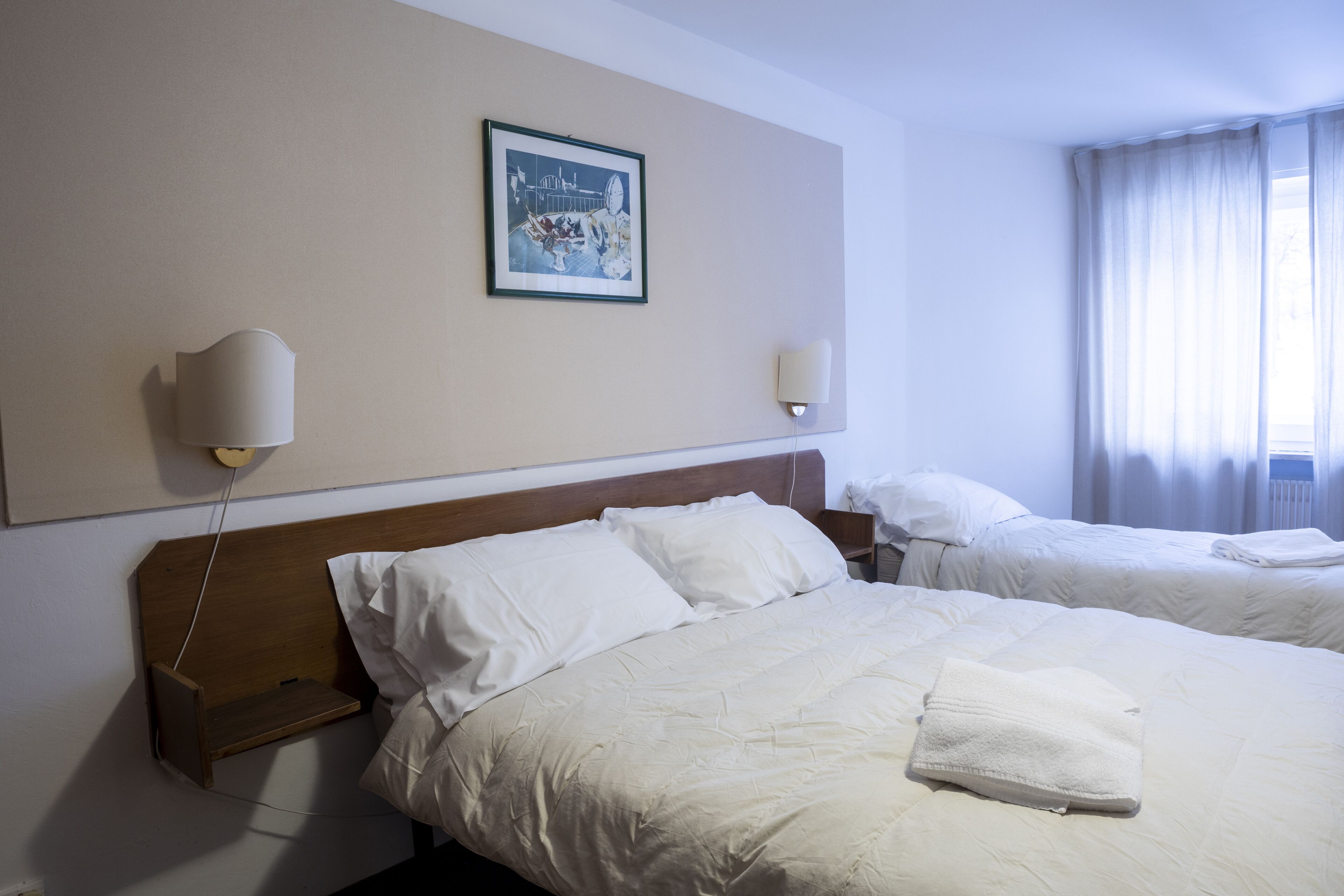 Hotellihuone, jossa on kaksi sänkyä, sängynpääty, yöpöytä, seinään kiinnitetty valaisin ja kehystetty kuva.