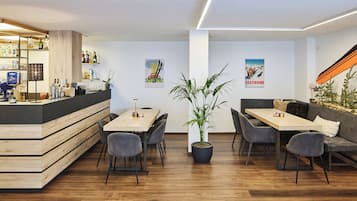A modern bar area with a wooden counter, seating with grey chairs, a central potted plant, and wall art.