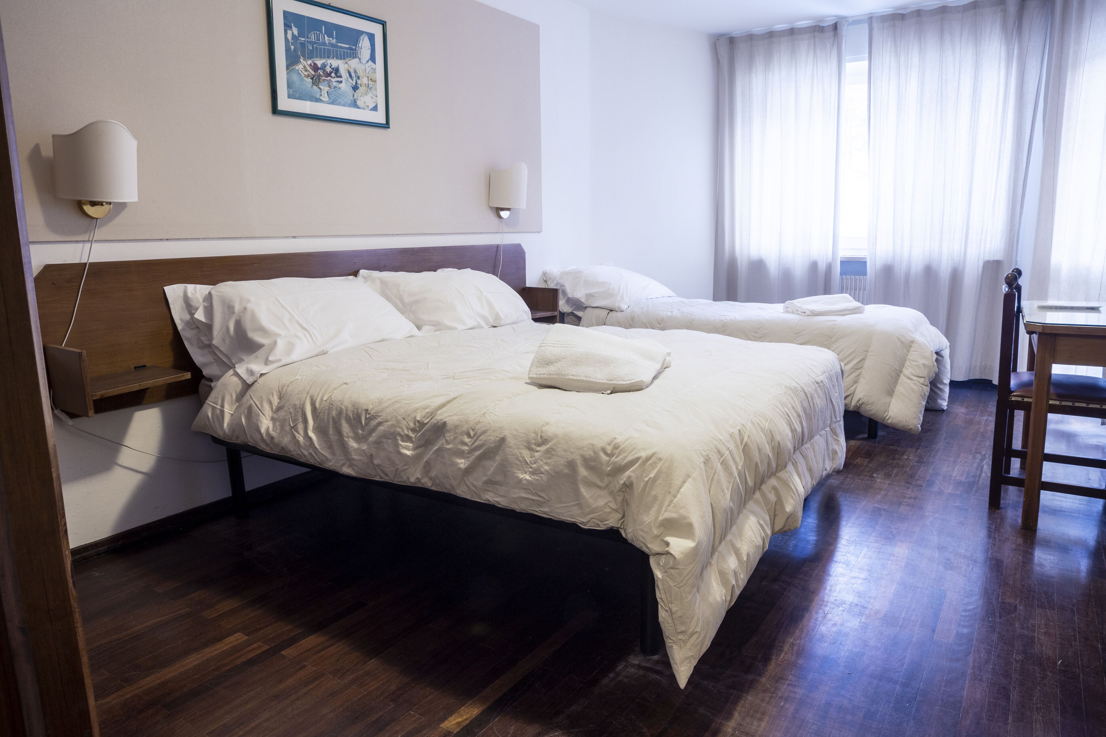 A hotel room with two beds, wooden headboards, a desk, and a chair.